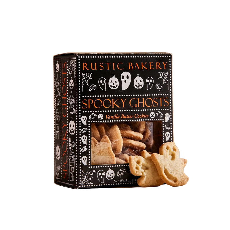 A box of Rustic Bakery Spooky Ghosts vanilla butter cookies sits on a light beige surface against a warm beige background. The black packaging features white Halloween-themed illustrations, and two ghost-shaped cookies are placed in front of the box.