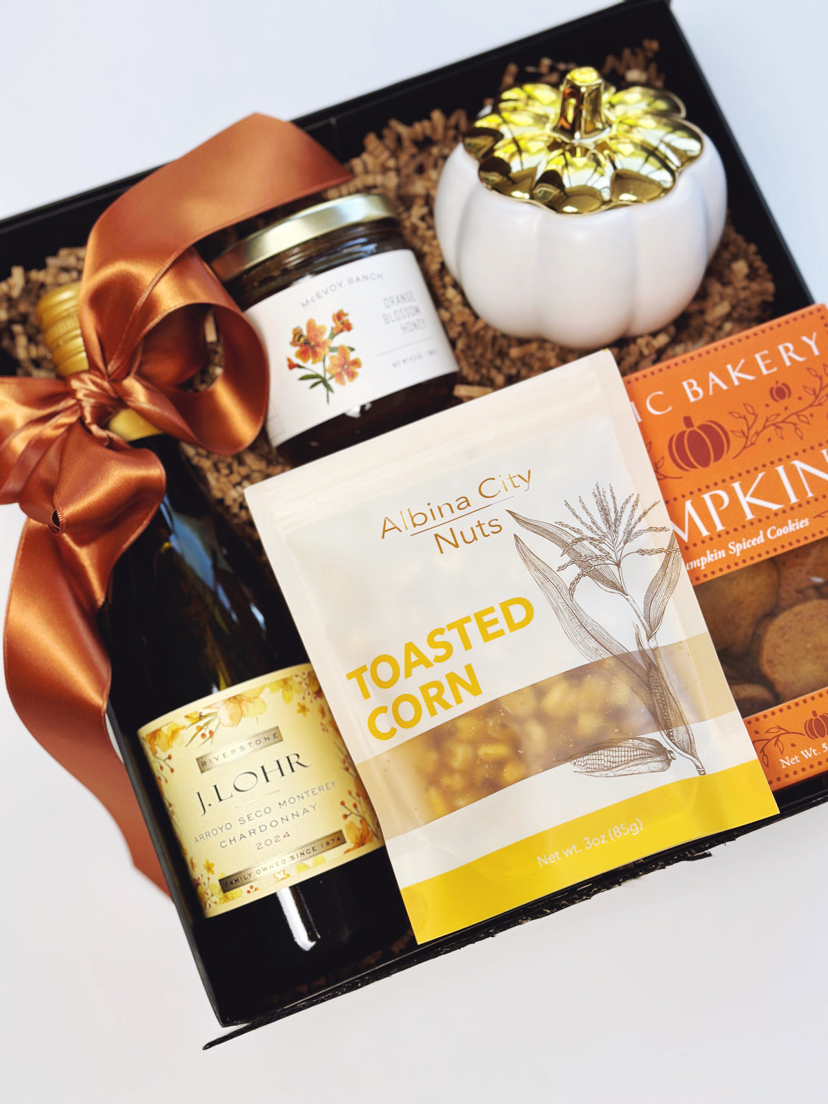 An autumn-themed gift basket set from with Albina Roasted Corn, featuring a bottle of chardonnay, a jar of honey, pumpkin cookies, a small white pumpkin Candle, and a Rust Colored ribbon, all arranged in a black box with shredded paper around the items.