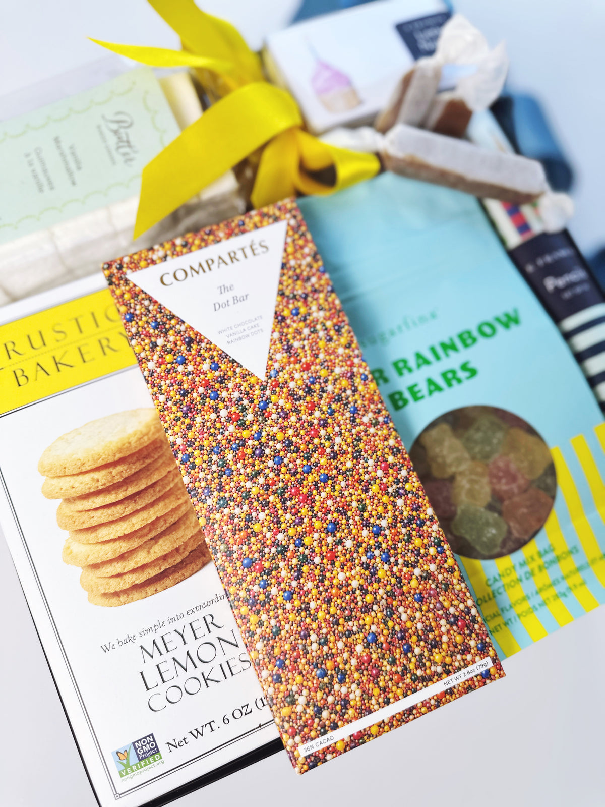 A gift basket arrangement featuring a Compartes 'The Dot Bar' in the center, surrounded by other packaged snacks and treats including a box of Rustic Bakers Meyer Lemon Cookies and a bag of gourmet rainbow gummy bears on a white background.
