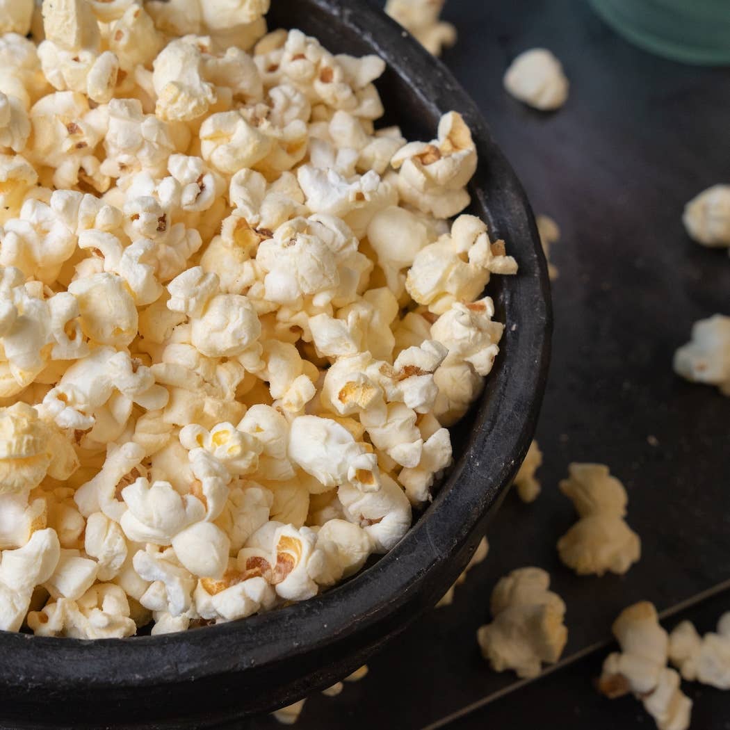 A close-up of fluffy white popcorn filling a dark bowl, with a few popcorn pieces scattered on a dark surface.