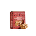 A box of Rustic Bakery Maple Leaves sugar cookies sits on a light beige surface against a warm beige background, with two leaf-shaped cookies placed in front of the package.